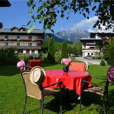 Ein gemütlicher Garten mit einem runden Tisch und Stühlen. Im Hintergrund sind Gebäude und Berge sichtbar, umgeben von üppigem Grün.