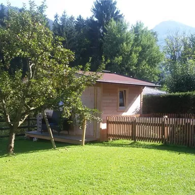 Ein schöner Garten mit einem Holzhaus und einem saftigen grünen Rasen. Im Hintergrund sind Bäume und Berge zu sehen.