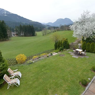 Ein gepflegter Garten mit Rasen, Blumen und einen schönen Blick auf die Berge. Zwei Gartenstühle laden zum Entspannen ein.
