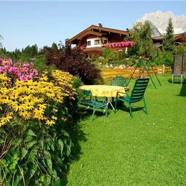 Ein schöner Garten mit bunten Blumen und grünen Stühlen. Im Hintergrund sind Häuser und Berge sichtbar.