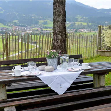 A cozy table outdoors, set with dishes and a bouquet of flowers. In the background, green meadows and mountains can be seen.