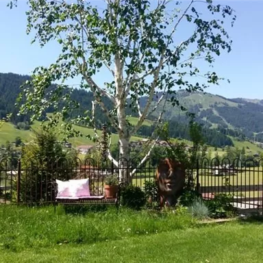 Eine malerische Landschaft mit Bergen im Hintergrund und grünem Gras. Ein schöner Baum steht in einem gepflegten Garten mit einem Zaun.
