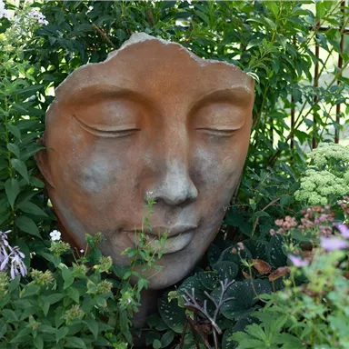 Ein Gesicht aus Ton, das aus einem Gartenbeet herausragt. Umgeben von bunten Blumen und Grünpflanzen, strahlt es Ruhe und Harmonie aus.