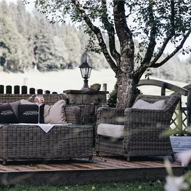 A cozy terrace with a rattan furniture set and cushions. In the background, green meadows and trees can be seen.