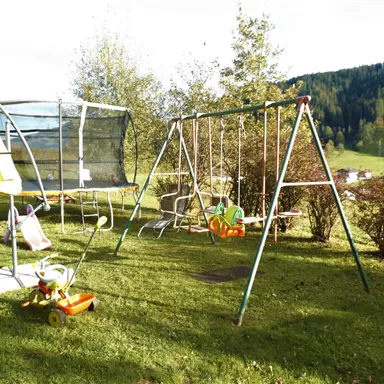 Ein Spielplatz im Garten mit einer Schaukel und einem Trampolin. Im Hintergrund sind Bäume und eine grüne Wiese zu sehen.