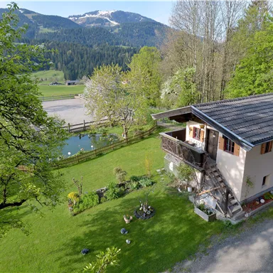 A picturesque house with a balcony, surrounded by lush greenery and trees. In the background, mountains and a tranquil pond extend.