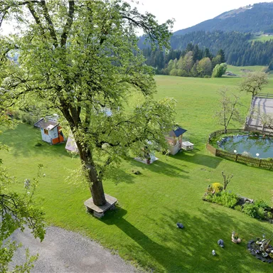 A peaceful landscape with a large tree, meadows, and small cottages. In the background, there is a pond and gentle hills.