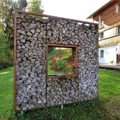 A decorative woodpile with a rectangular window in which flowers are growing. In the background, a modern wooden house is visible.