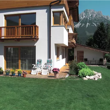 A modern house with a well-maintained garden and cozy loungers on the terrace. In the background, majestic mountains can be seen.
