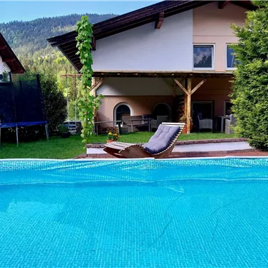 Ein schöner Pool mit klarem Wasser und einer Liege daneben. Im Hintergrund steht ein Haus mit einem überdachten Bereich und viel Grün.