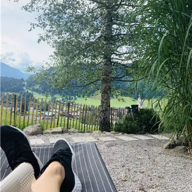 Eine entspannte Liegeposition in der Natur mit einem Blick auf grüne Wiesen und Berge. Ein Baum und hohe Pflanzen umrahmen die Szene.