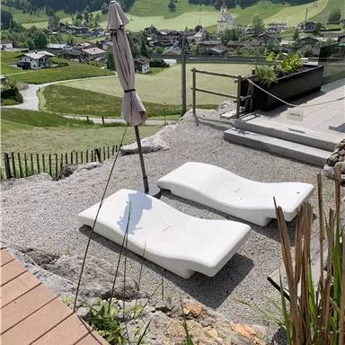 Zwei Liegen stehen auf einem Kiesplatz mit Blick auf eine malerische Landschaft. Im Hintergrund sind grüne Hügel und ein Dorf zu sehen.