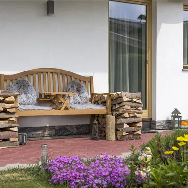 Eine gemütliche Bank mit Kissen, umgeben von einem gepflegten Garten. Neben der Bank liegt ein Holzstapel und bunte Blumen blühen.