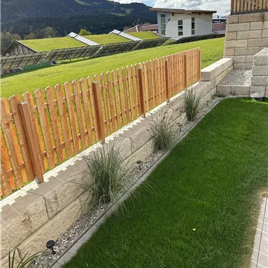 A well-maintained garden with green grass and a wooden picket fence. In the background, mountains and buildings are visible.