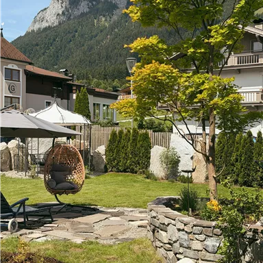 A beautiful garden with a hammock, well-kept lawn, and stone areas. In the background, mountains and buildings are visible.