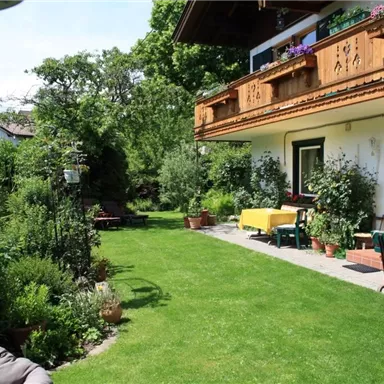 A well-maintained garden with green grass and many plants. In the background, there is a traditional house with a balcony.