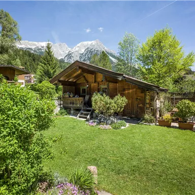 A rustic wooden house in a green garden, surrounded by trees and flowers. In the background, snow-covered mountains can be seen.