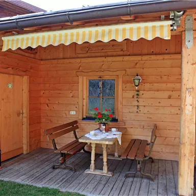Ein einladender Außenbereich mit einer Holzveranda und einem kleinen Tisch. Zwei Bänke und eine Markise sorgen für Gemütlichkeit.