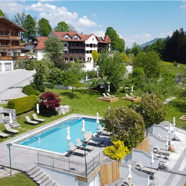 A luxurious hotel with a swimming pool and a well-kept garden. In the background, mountains and a clear blue sky can be seen.