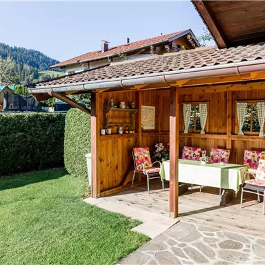 A cozy outdoor area with a wooden house and a table set with flower chairs. In the background, there is a green meadow and mountains.