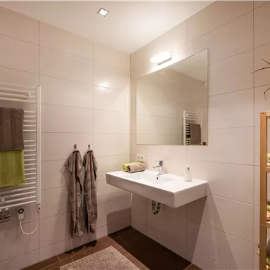 A modern bathroom with bright tiles and a large mirror. Towels and a shelf with grooming products complete the decor.