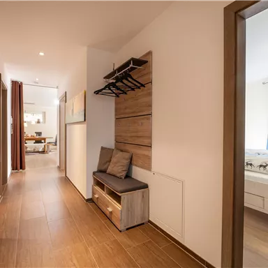 A bright hallway with wooden flooring and a seating area. On the left side, there are coat hooks and access to a living room.