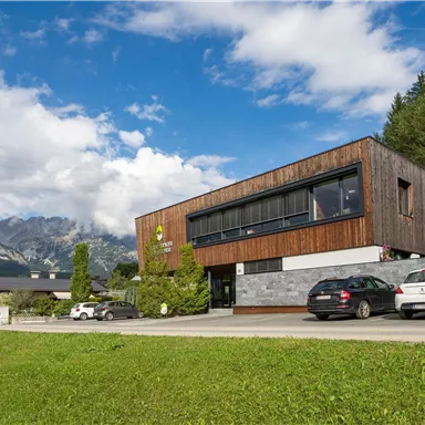 A modern building with wooden elements and large windows stands in a picturesque landscape. In the background, mountains and a clear sky can be seen.