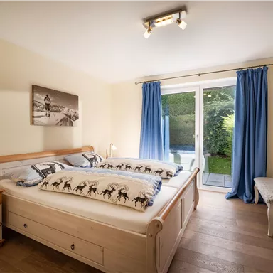 A bright bedroom with a large double bed and wooden furniture. Blue curtains hang in front of the windows, and there is a view outside into the garden.