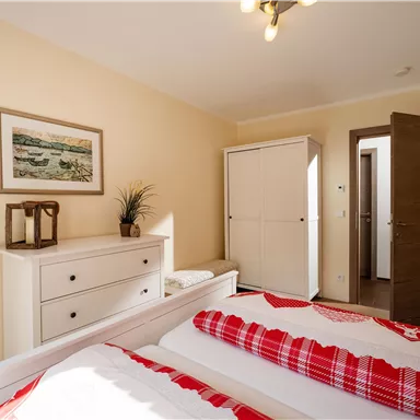 A bright bedroom with two single beds and a red-and-white blanket. A picture hangs on the wall, and there is a white wardrobe as well as a wooden chest of drawers.