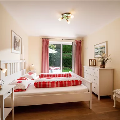 A cozy bedroom with a double bed, red decorative pillows, and a desk. Large windows provide ample daylight and a view of the greenery.