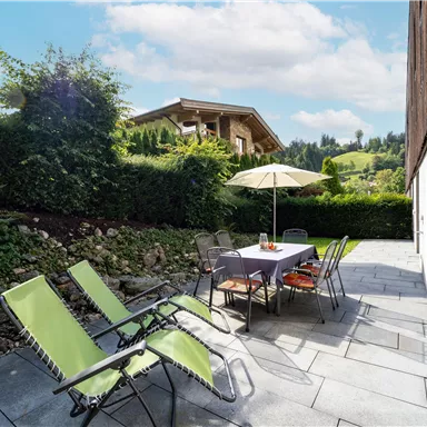 An inviting patio with a table and chairs, surrounded by green plants. In the background, there is a wooden house and a hilly landscape.