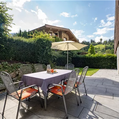 A cozy outdoor seating area with a table and chairs under a sunshade. In the background, green hills and an attractive house can be seen.