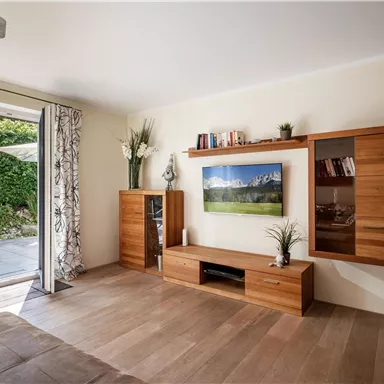 A modern living room with wood-colored furniture and a large window. In the background, a green garden landscape can be seen.