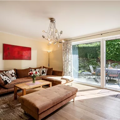 A bright living space with a cozy sofa and a wooden table. Large windows offer a view of a garden area.