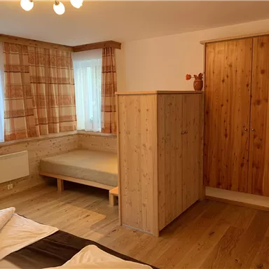 A simple bedroom with wooden furniture and a bed. The walls are light and there are large windows with curtains.