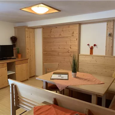 A cozy living space with wooden furniture and a table. There are vases with flowers on the shelf and a television is visible.