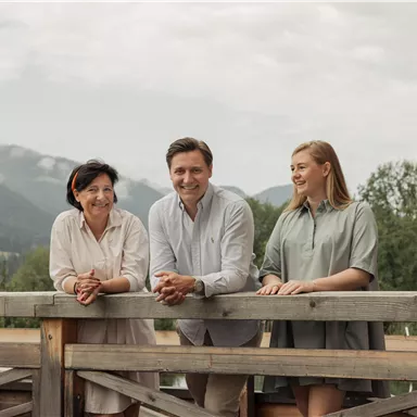 Eine Gruppe von drei Personen steht auf einer Holzplattform mit Blick auf die Berge. Die Szene vermittelt eine freundliche und entspannte Atmosphäre.