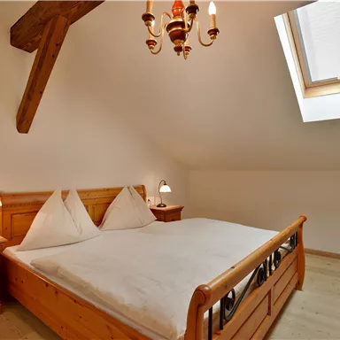 A cozy bedroom with a large bed and wooden furniture. The room has a bright atmosphere thanks to a window and a ceiling lamp.