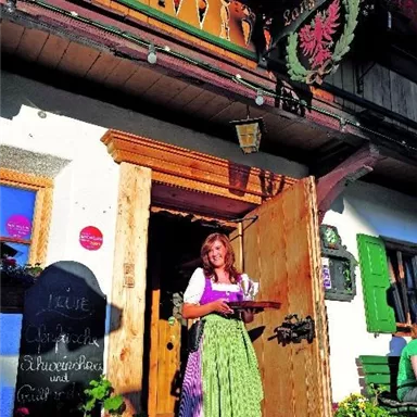 A cheerful woman in traditional attire holds a tray in front of a cozy inn. The facade of the building is decorated with flowers and an inviting atmosphere.