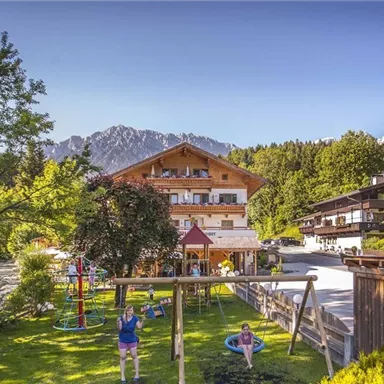 Ein malerisches Bergdorf mit traditionellen Holzhäusern und grünen Wiesen. Im Vordergrund gibt es einen Spielplatz und die beeindruckenden Berge im Hintergrund.
