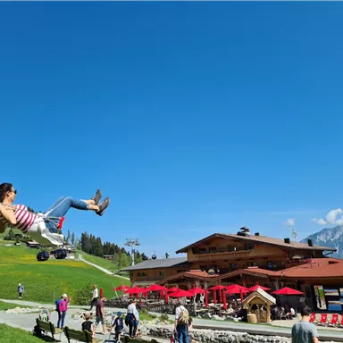 A woman swings on a swing in front of a mountain restaurant. In the background, green meadows and a blue sky can be seen.