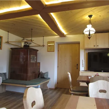 Ein gemütlicher Raum mit Holzdecke und einem traditionellen Kachelofen. Es gibt einen Tisch mit Stühlen und einen Fernseher an der Wand.