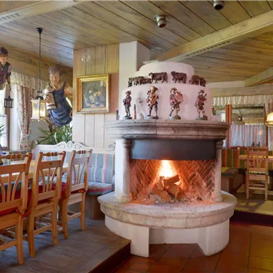 A cozy restaurant with wood decor and an open fireplace. In the background, tables and chairs as well as decorative figures are visible.