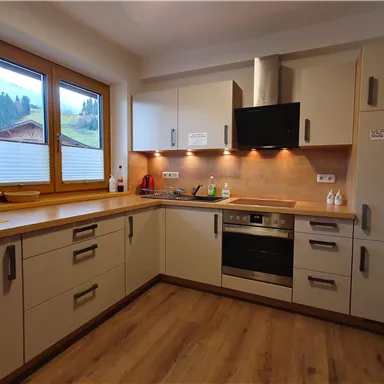 A modern kitchen with wooden details and a large window. The equipment includes a stove, a refrigerator, and various kitchen appliances.