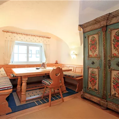 A cozy dining area with a wooden table set and a colorful, painted chest of drawers. The walls are light, and the light falls gently through the window.