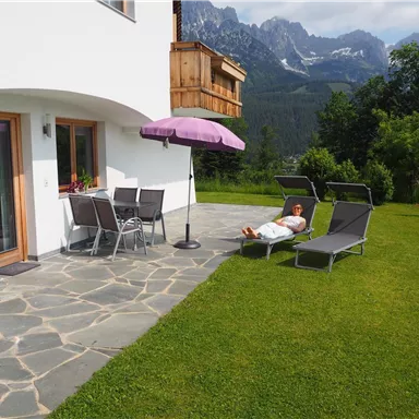 Ein geräumiger Außenbereich mit Liegen und einem Tisch unter einem Sonnenschirm. Im Hintergrund sind grüne Wiesen und Berge sichtbar.