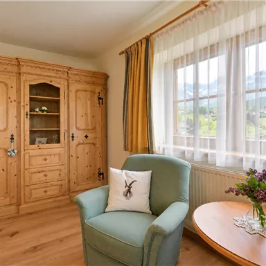 Ein gemütliches Wohnzimmer mit einerm blauen Sessel und einem Holzschrank. Große Fenster lassen viel Licht herein und bieten einen Blick auf die Berge.