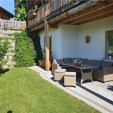 Eine gemütliche Terrasse mit einem grauen Sitzbereich und einem Tisch. Der Garten ist grün mit Sträuchern und einer Treppe im Hintergrund.