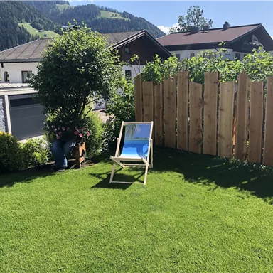 Ein schöner Garten mit grünem Rasen und einer Liege. Im Hintergrund sind Holzverkleidungen und einige Bäume zu sehen.