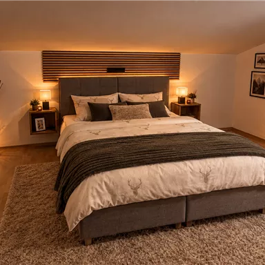 Ein gemütliches Schlafzimmer mit einem großen Bett und warmem Licht. Dekorative Elemente wie Pflanzen und Bilder sorgen für eine einladende Atmosphäre.
